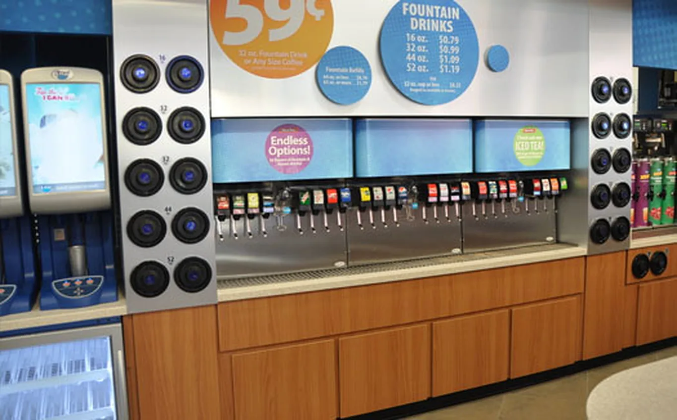 A convenience store fountain drink station with a row of soda dispensers, cup holders and a fountain drinks price board.