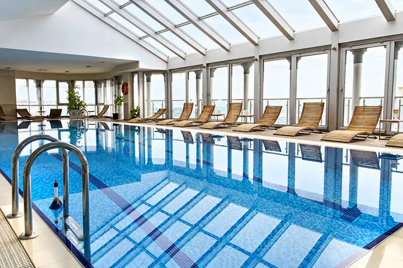 An indoor commercial swimming pool under a glass roof, with lounge chairs along the windows.