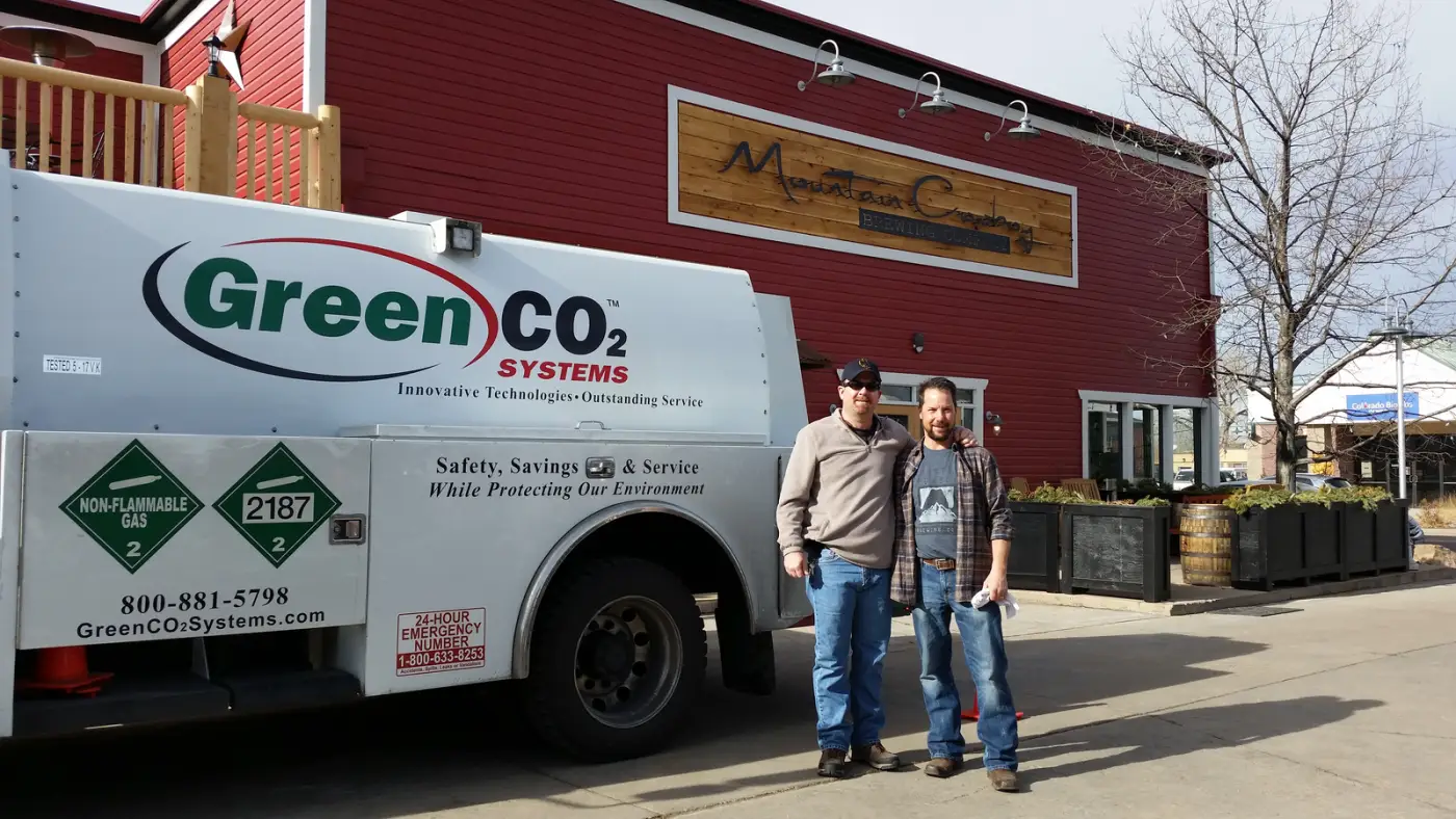 A Green CO2 Systems bulk delivery truck parked outside Mountain Cowboy Brewing on its grand opening day, with two men standing beside it.